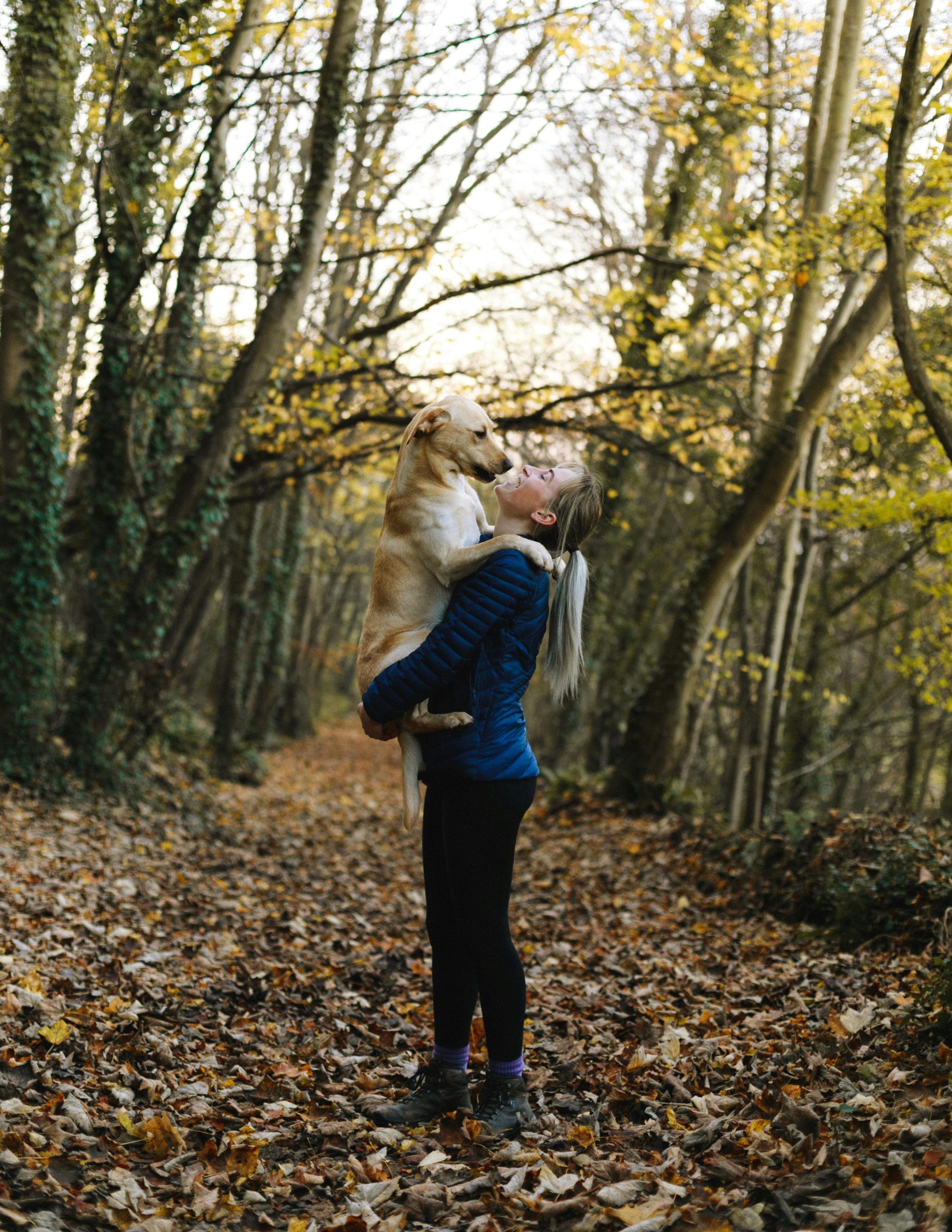 Person standing on leaf-covered path in forest holding a light-colored dog.