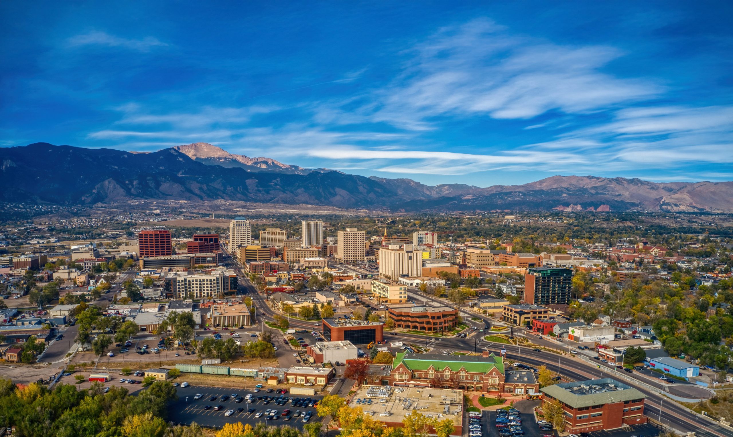 Aerial view of downtown Colorado Springs with modern buildings, green spaces, and mountains in the background under a blue sky with wispy clouds.