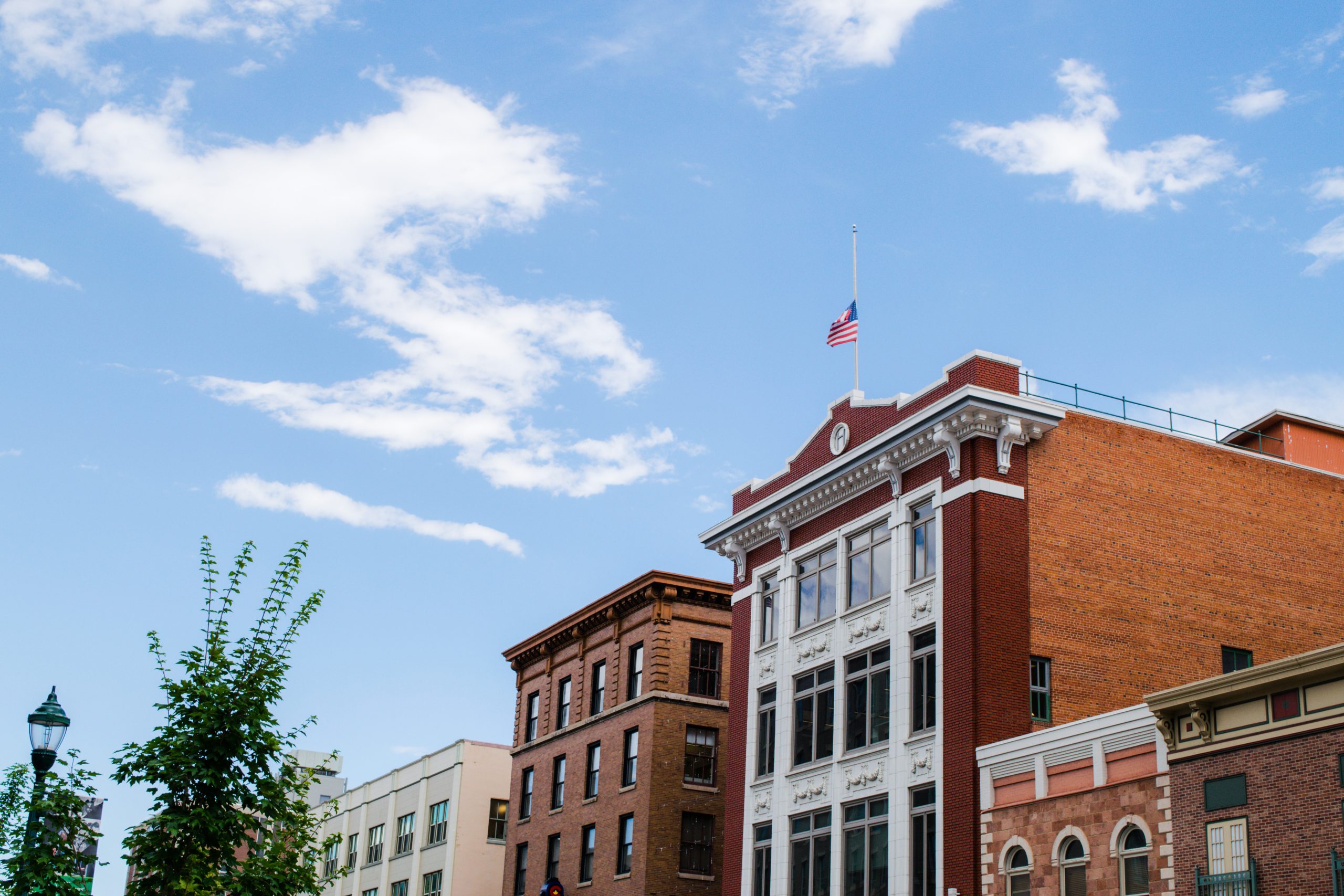 A row of historic brick buildings under a blue sky with scattered clouds. An American flag flies at half-staff atop one of the buildings. A leafy tree and streetlamp are visible on the left.
