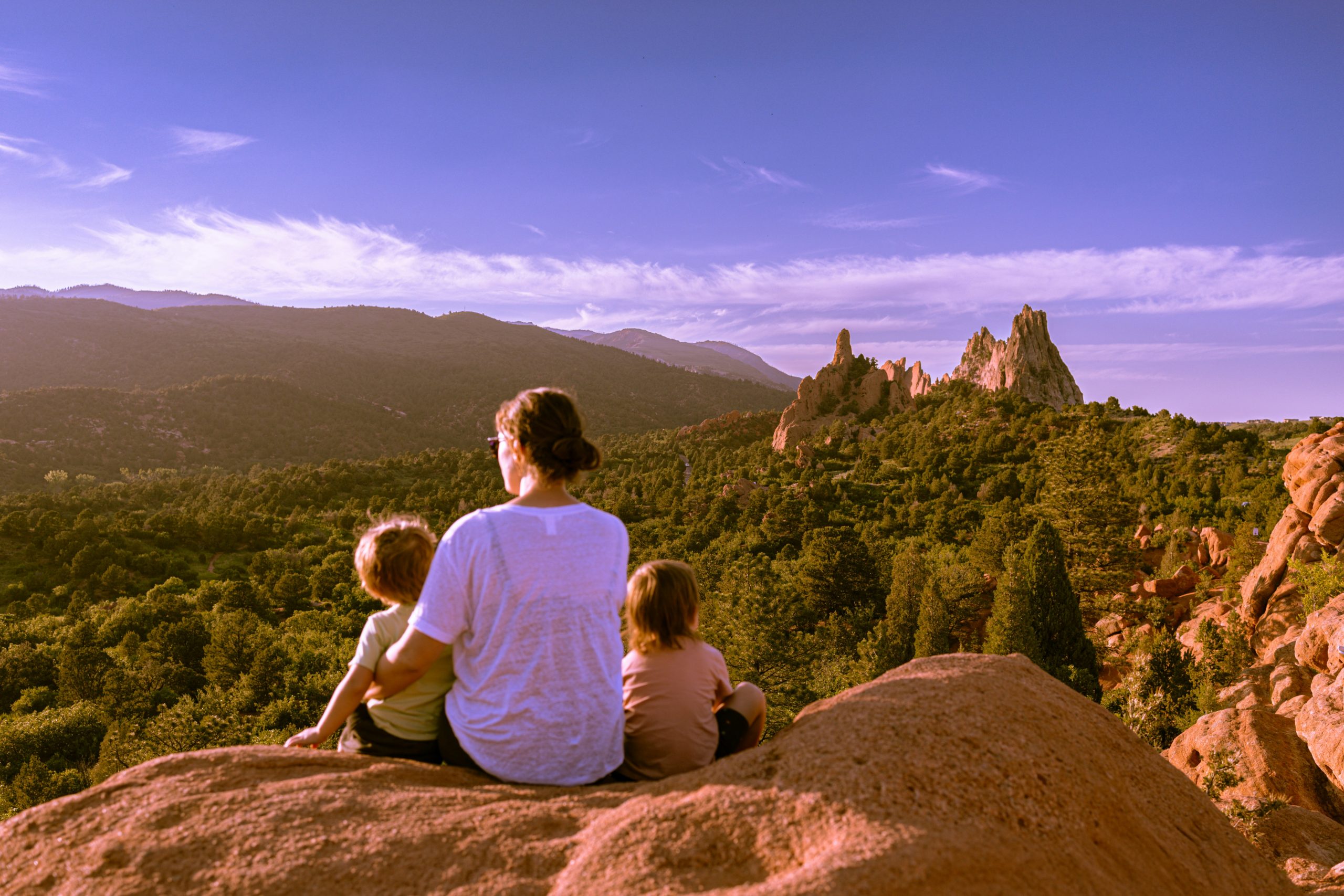 A woman sits on a rocky ledge with two children, overlooking a lush green valley and dramatic rock formations under a clear blue sky with mountains in the distance.