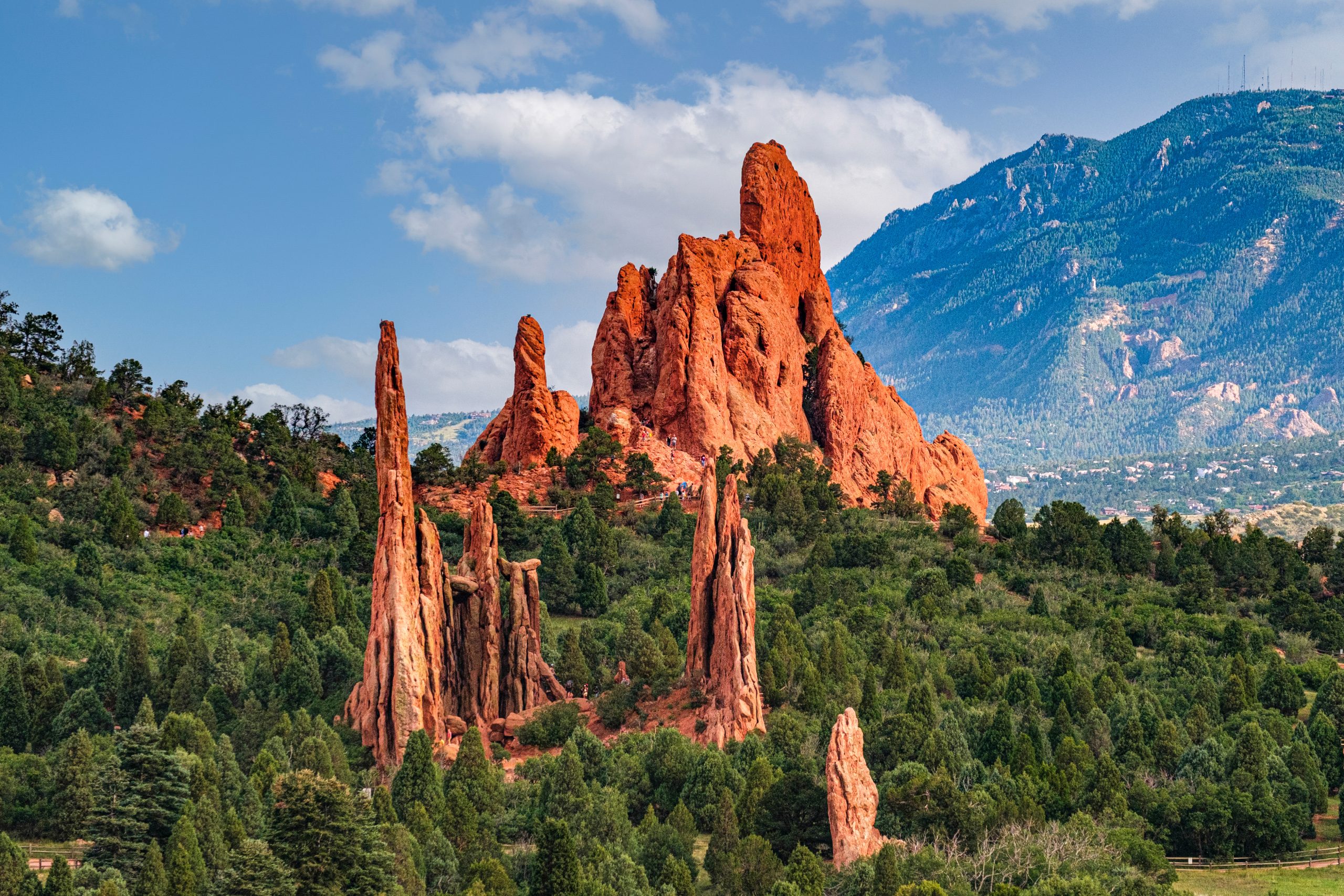 Tall, jagged red rock formations rise from a green forested landscape, with a mountain range and blue sky with scattered clouds in the background.