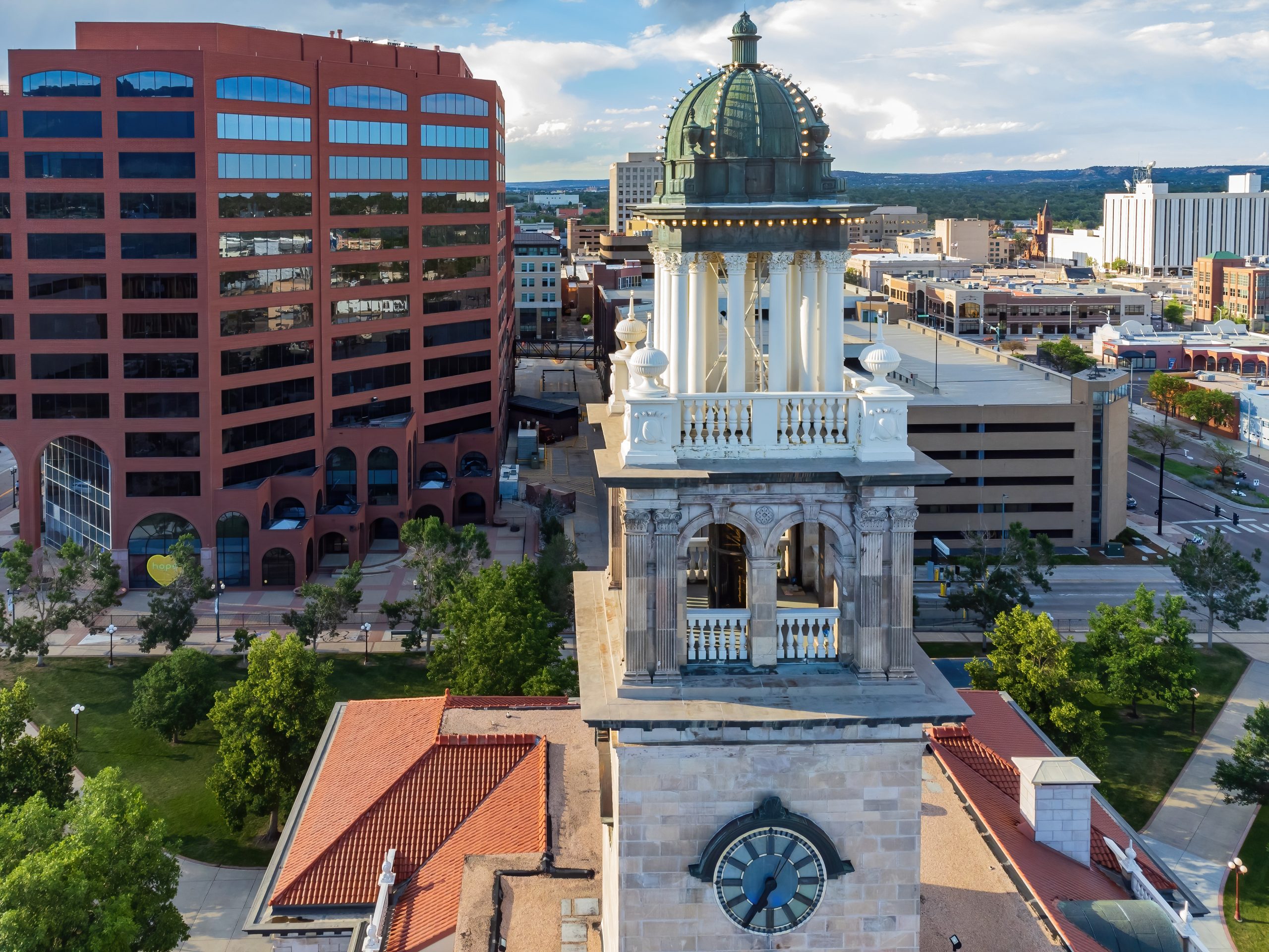 Aerial view of a clock tower with a domed roof, surrounded by red-roofed buildings and modern offices in a cityscape under a partly cloudy sky.