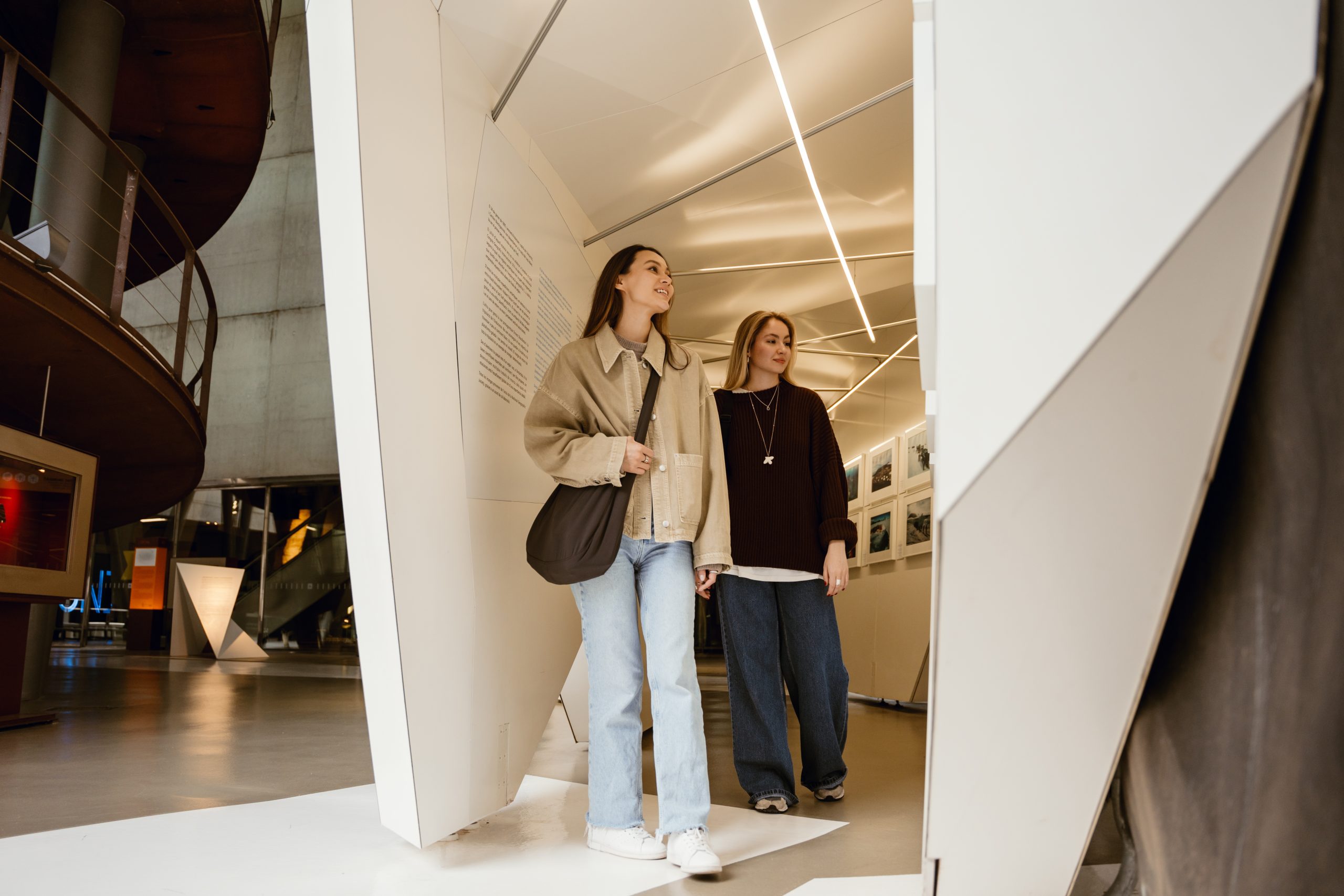 Two young women walking through a modern, well-lit hallway or exhibit space, looking around and smiling. Both are casually dressed and the interior features geometric white panels and photos on the wall.